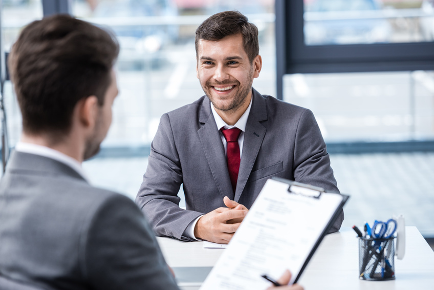 Smiling young businessman looking at manager with clipboard at job interview